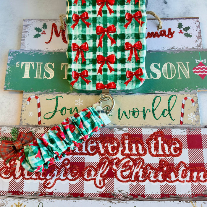 Plaid Tidings & Bows scrunchie phone wrist strap clipped below the matching gingham bow iPhone case on wooden Christmas signs