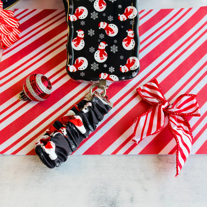 Snowman scrunchie phone wrist strap clipped to the matching snowman Wavy Luxe iPhone case, on candy cane stripes with a Santa hat and ornaments