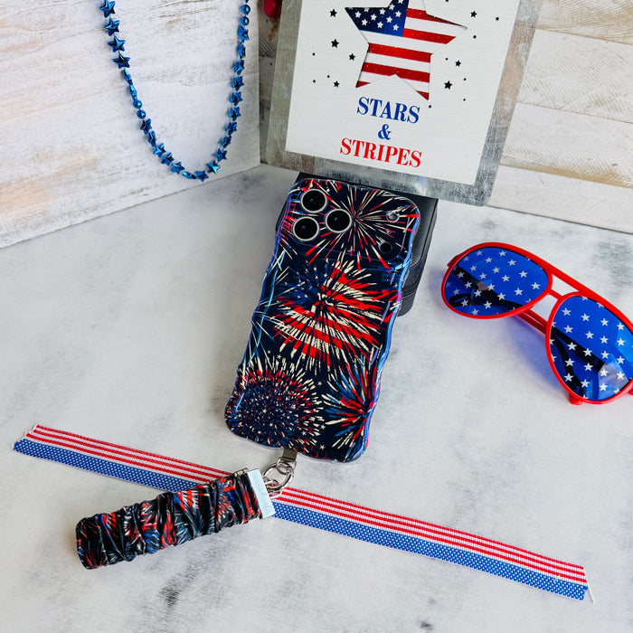 USA Fireworks scrunchie phone wrist strap laid out with the matching firework print Wavy Luxe iPhone case, star sunglasses and a red white and blue beaded strand