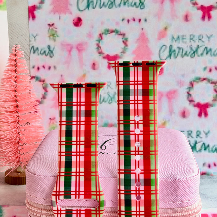Both straps of the Merry Little Plaid silicone Apple Watch band standing on a pink box beside a pink bottle-brush tree