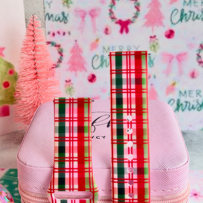 Both straps of the Merry Little Plaid silicone Samsung Galaxy Watch band standing on a pink box beside a pink bottle-brush tree