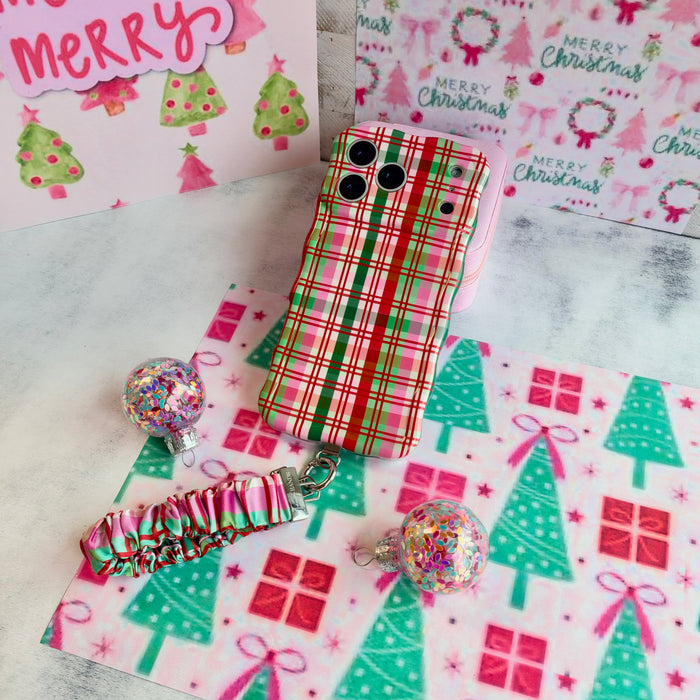 Merry Little Plaid iPhone case with the matching scrunchie phone wrist strap attached, laid flat on pink and mint Christmas paper