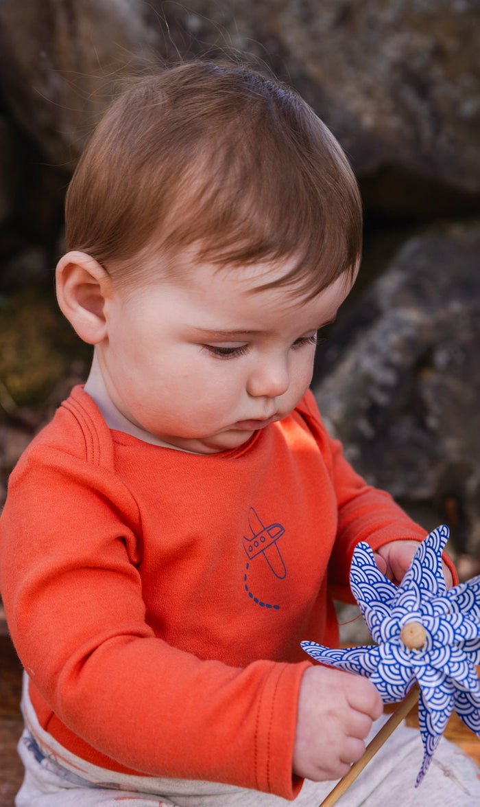 Baby wearing the Deux par Deux organic cotton onesie in coral with airplane detail, view 2