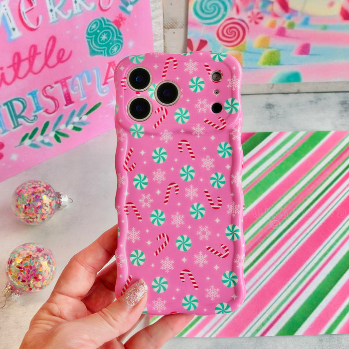 Hand holding the Peppermint Cutie Wavy Luxe MagSafe iPhone case, bubblegum pink with mint peppermints and candy canes, by a Merry Little Christmas sign
