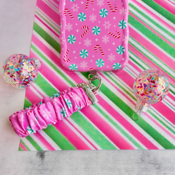 Peppermint Cutie scrunchie phone wrist strap clipped to the matching pink peppermint iPhone case, on candy stripe paper with glitter ornaments