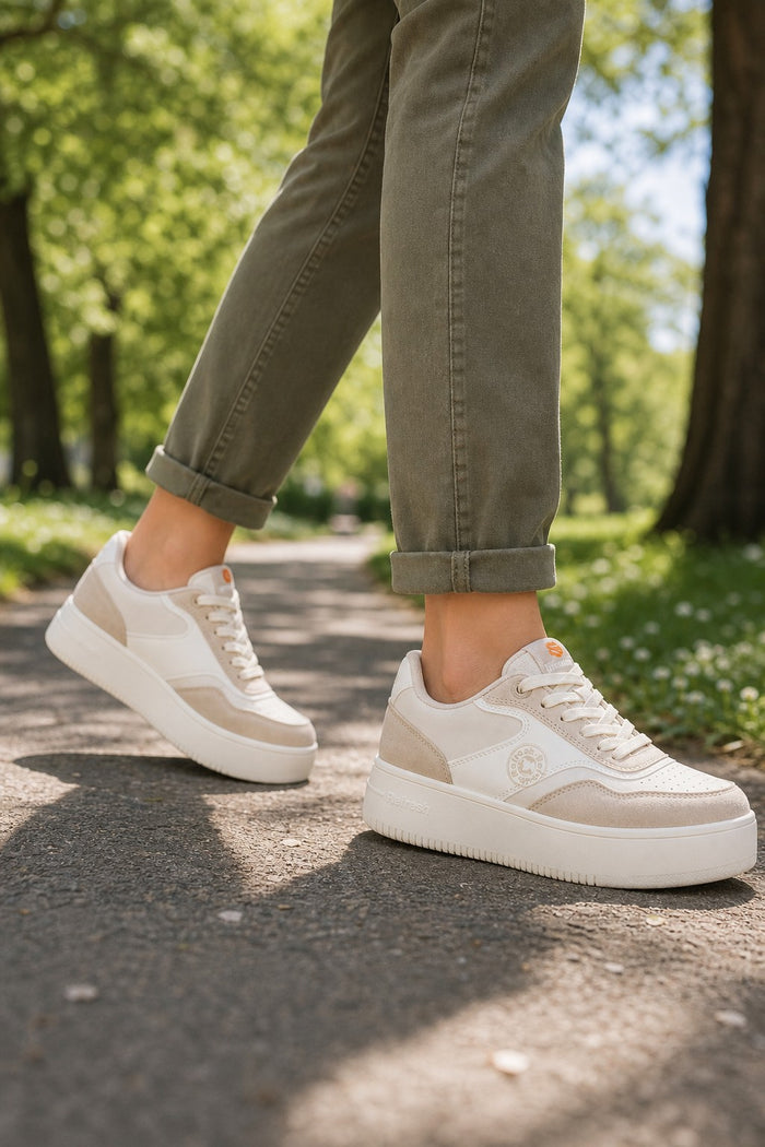 A person wearing beige and white platform sneakers with olive green cuffed pants, walking on a sunlit paved path in a park.
