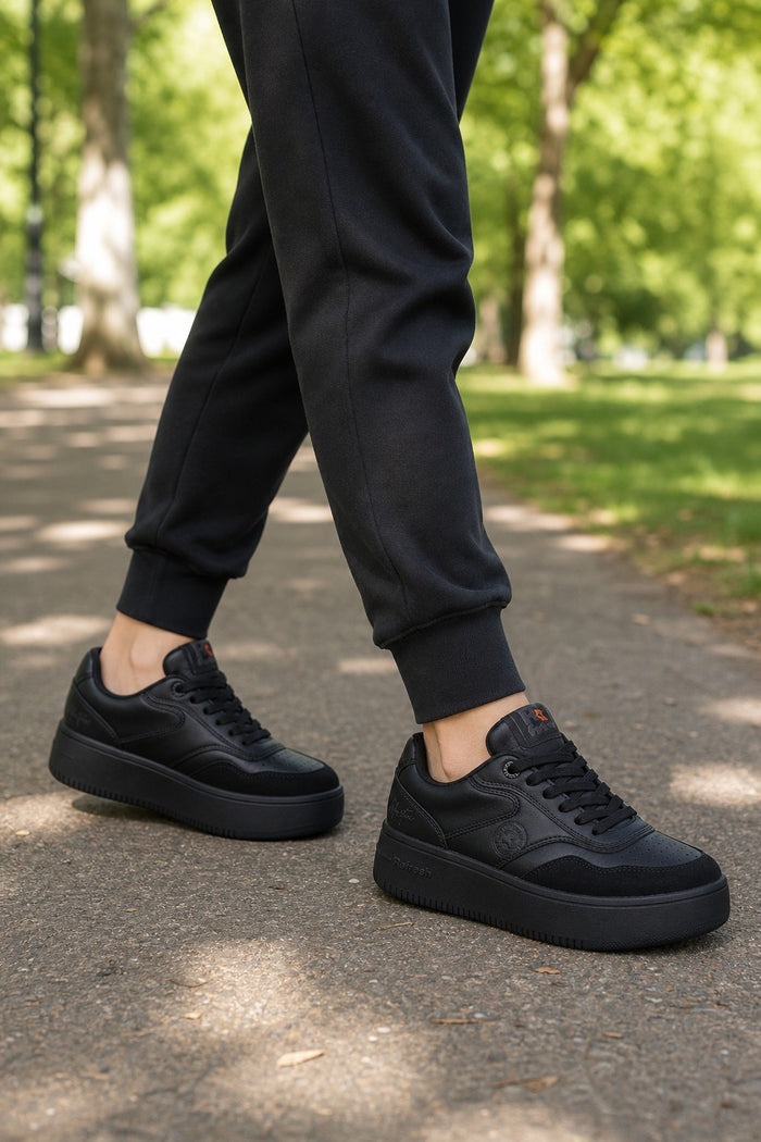 A person wearing black platform sneakers and black jogger pants walking on a paved path in a sunny park with green trees in the background.