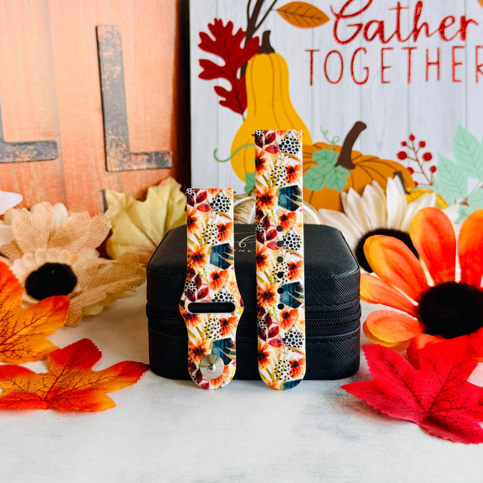 Sienna Petals Samsung Galaxy Watch band shown in both size options on a black stand against an autumn leaf and sunflower backdrop.