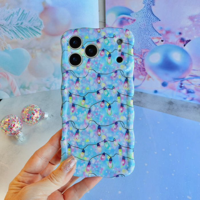 Hand holding the Twinkle All The Way Wavy Luxe MagSafe iPhone case, strands of pink, violet, aqua and lime Christmas lights on pastel blue