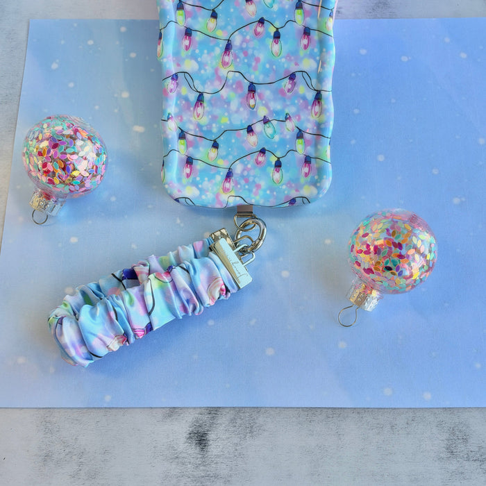 Twinkle All The Way scrunchie phone wrist strap clipped below the matching Christmas lights iPhone case between two glitter ornaments