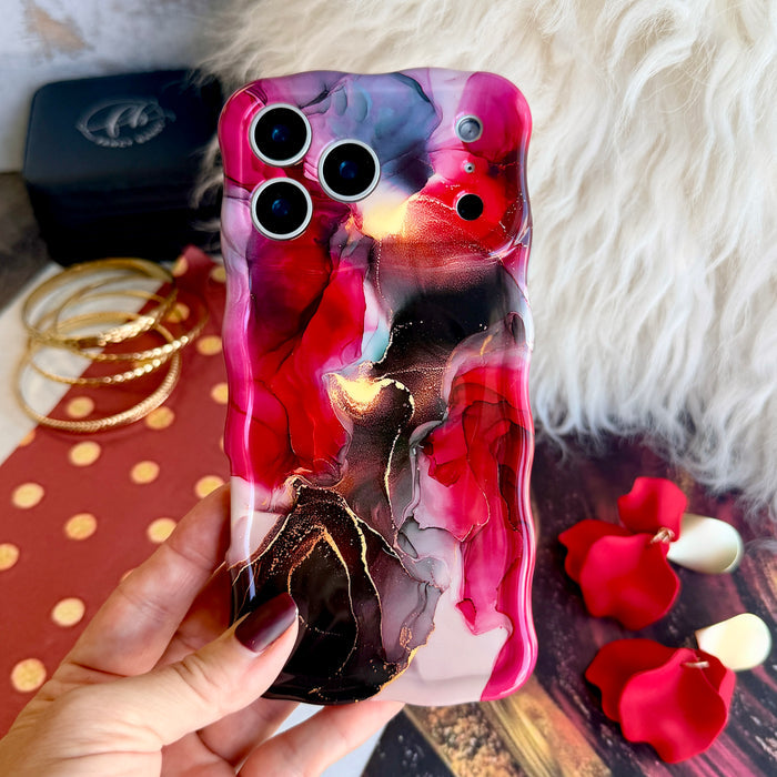 Hand holding the Velvet Vixen crimson and black alcohol-ink marble Wavy Luxe iPhone case over white fur, with stacked gold bangles and red rose petals.
