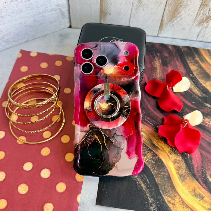The Velvet Vixen marble iPhone case wearing the matching crimson and gold MagSafe grip ring propped as a kickstand, styled with gold bangles and red rose petals.
