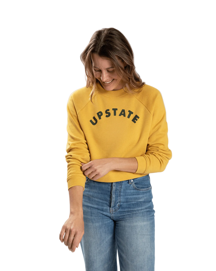 Woman wearing a yellow 'UPSTATE' sweatshirt and blue jeans on a white background